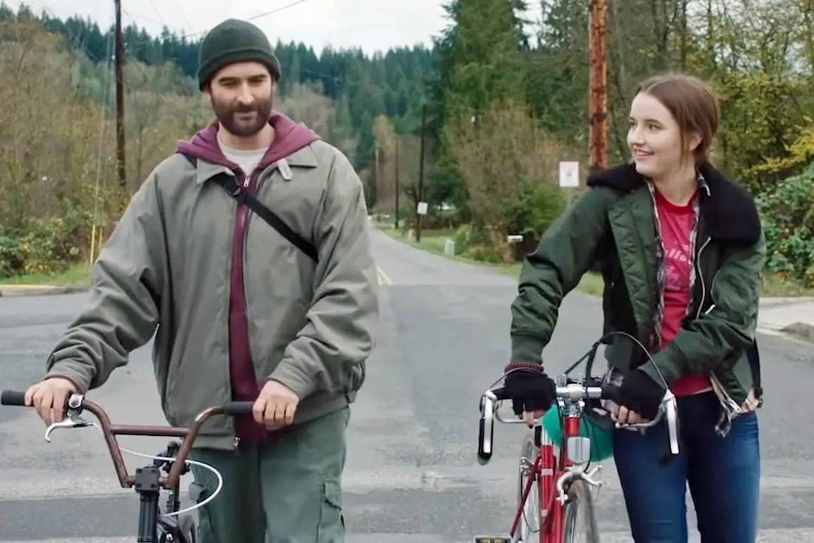 Photo from the 2017 movie Outside In. Jay Duplass and Kaitlyn Dever are walking their bikes wearing winter clothes on a road with tall trees on hills behind them. Dever has a 10 speed and is smiling as she looks at Duplass, who has a BMX bike.